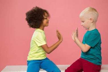 Küçük çocukların yan görüntüsü. Mutlu beyaz çift ve Afrikalı Amerikalı çocuklar gülümsüyor, yakınlarda oturuyor, oynuyorlar. Pembe arka planda izole edilmiş.. 