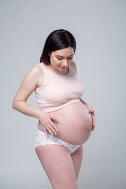 İç çamaşırlı, hamile, esmer bir kadın. Duygusal stüdyo fotoğrafı, beyaz bir arka planda hamile. Hamile bir kadının göbeği yuvarlak. Hamileliğin son ayı. Bebeği bekliyorduk. Bej renkler. Göz teması.