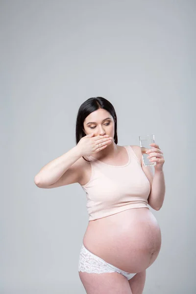 Pregnant caucasian woman in underwear with a glass of water in hand. Emotional studio photo pregnancy on a white background. To drink or not to drink water? Last month of pregnancy. Looks at a glass with a drink. Health Care Concept