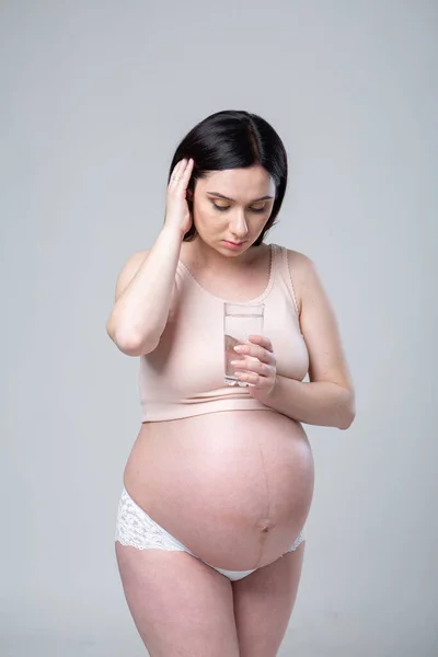 Pregnant caucasian woman in underwear with a glass of water in hand. Emotional studio photo pregnancy on a white background. To drink or not to drink water? Last month of pregnancy. Looks at a glass with a drink. Health Care Concept