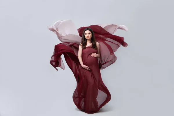Güzel hamile kadın. Burgonya ve pembeden uçan kumaşlar giymiş bir kız. Hamile bir kadının zarif duruşu, elleriyle karnını nazikçe tutuyor. Uzun saçlı. Gri arkaplan, stüdyo ışığı.