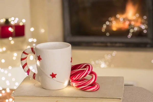 Large white mug with a warm drink, sweets, book. Fireplace as background. Gifts, holiday atmosphere, expectation of magic, new year, christmas concept. Warm cozy house. Nobody. Horizontal. Copy space