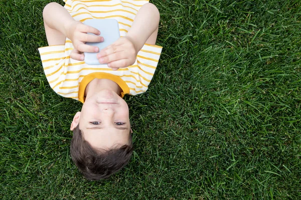 Portrait of a cute child. Flipped upside down, lies on the grass. Smiles. Holding a smartphone in his hands. Eye contact. Copy space. Concept of internet, summer, holidays, childhood, applications.