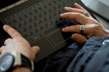 Erkek eller siyah bir dizüstü bilgisayarda klavye kullanıyor. - Yakın çekim. Parmaklarda zarif çelik halkalar, saatler. Çevrimiçi iş kavramı, serbest çalışma, mesafe çalışması, internet üzerinden iletişim, aygıtlar.