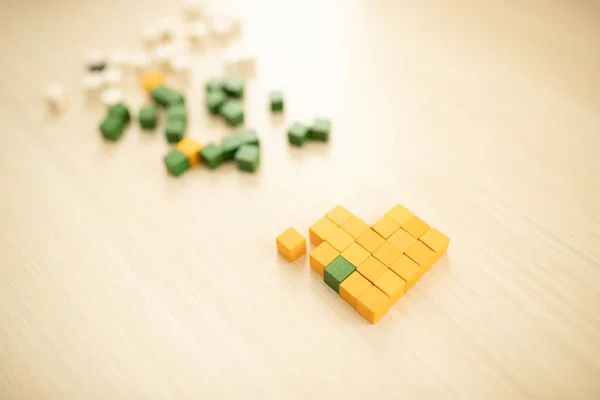 Heart made of wooden pixels rests on tabletop. Yellow cubes, one green - the symbol of the missing element. Problems of autistic people, teaching children with special needs concept. Selective focus.