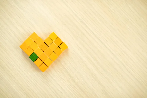 Heart made of wooden pixels lies on light wooden surface. Yellow cubes, one green - symbol of the missing element. Autistic people problem concept, care for education of children with special needs.