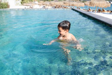 5-6 yaşındaki mutlu çocuk otel havuzunda oynuyor. Yüzüne su damlıyor. Gülümsüyor, gülüyor. Parlak, güneşli bir gün. Temiz su. Kafkasyalı. Yüzme konseptinin.