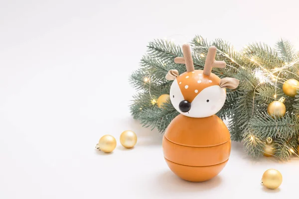 Altın toplar ve ışıklarla süslenmiş Noel ağacının yanındaki sevimli oyuncak geyik. Yeni yıl kartı. Beyaz arka plan. Çocukluk, mutlu tatil konsepti. Yatay olarak. Uzayı kopyala Tahta eko oyuncak.