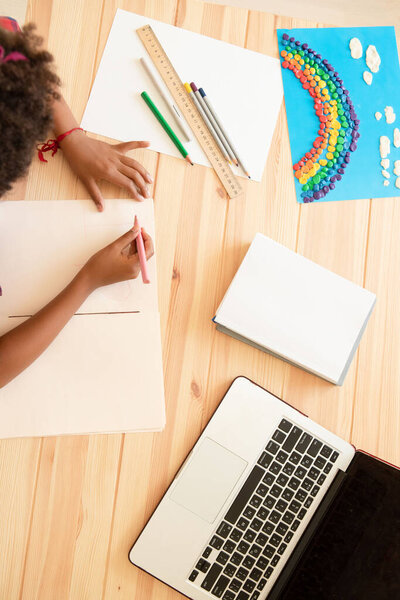 Ребенок смешанной расы рисует на столе. Рядом находится ноутбук, блокноты, карандаши, линейка и радуга ручной работы. Концепция дистанционного обучения, домашнее задание, возвращение в школу. Вид сверху. 