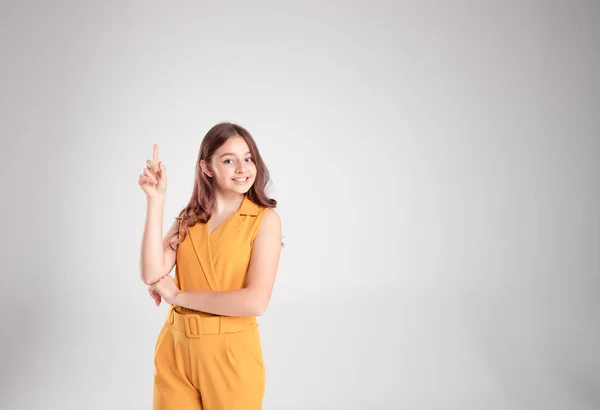 Güzel bir kız işaret parmağı havada bir jest gösterir. Diş tebessümü, sağlıklı cilt. Göz teması. Sarı giyinmiş. Stüdyo ışığı. Gri arka plan. Uzayı kopyala Yatay.