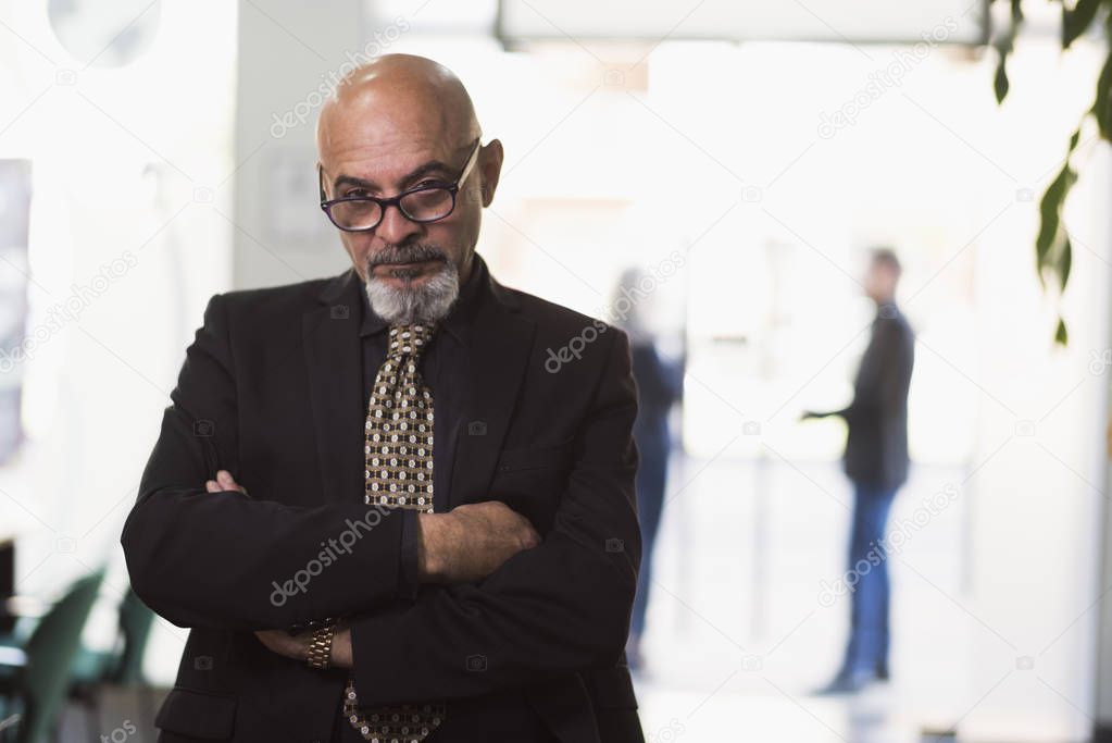 Mature boss in office posing and looking at camera with people in background windows