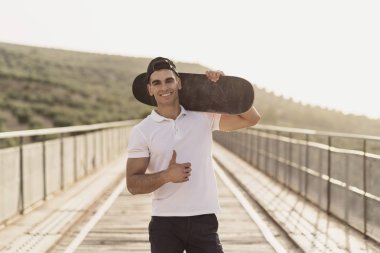 Kaykaylı genç adam köprüde gülümseyen kameraya bakıyor.