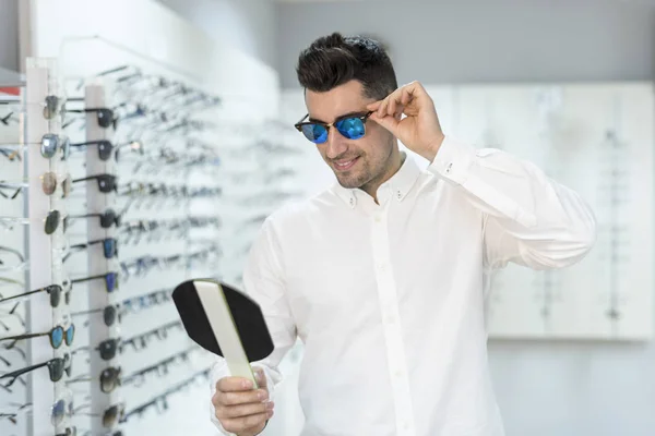 Человек примеряет солнечные очки в оптическом магазине, смотрит в зеркало, чтобы увидеть — стоковое фото