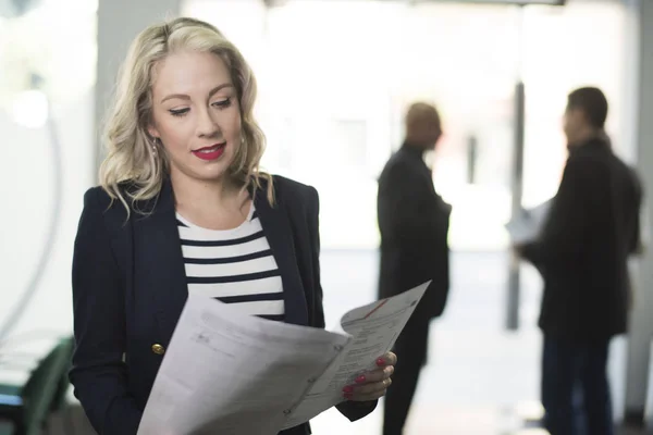 Blonde woman in office looking papers with people in the background