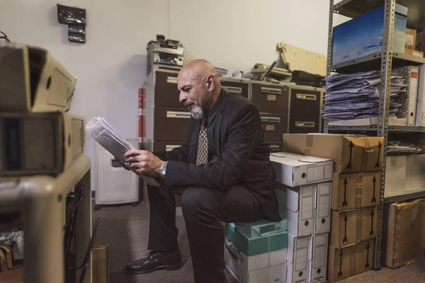 mature man in office file warehouse rests sitting on a box and checks documentation