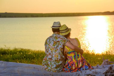 Kız adamla göl kıyısında oturuyor. Günbatımı su arka plan üzerinde. Parlak ve sulu yeşil çim ve yaz güneşin sıcak günbatımı. Aşık doğada kucaklamak. Şapka insanlar. Mutlu genç insanlar tatil.