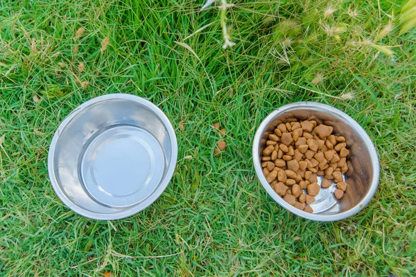 Bowls with food and water. Pet feeding cup. Metal plate for eating dogs. Dry useful balanced pet food. A cup with food and water stands on the green grass.