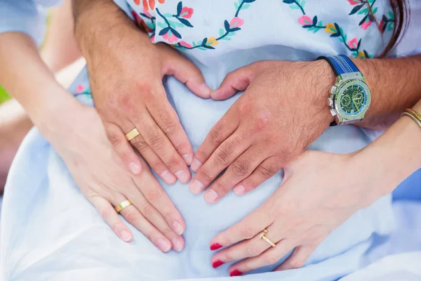 Nakış ile beyaz giysili bir kadına. Gebelik. Bir kalp şeklinde karnına elleri katlanmış. Olumlu duygular. Bir mucize bekliyorum. Nikah yüzükleri. Doğada piknik.