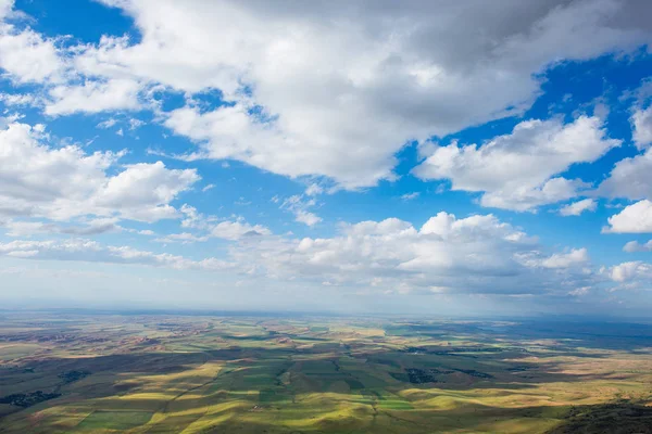 Blue sky with fluffy clouds. White cirrus clouds illuminated by the sun. Greenery small hills on the plateau. Earth meets the sky. Earth aerial view. Horizon away. Beautiful plain under the clouds.