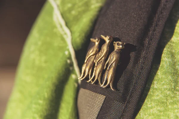 Brooch, on which there are three mongooses. Icon with animals. Backpack strap on the background of a green jacket. Metal badge on the strap from the backpack. Mongoose.