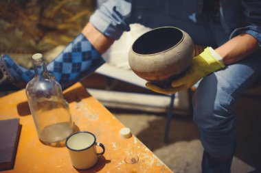 Bir adam bir demir pota ocağı için tutar. Adam elinde. , Atölye. Metal ile çalışır. Demirci cam şişe. Demir kupa. Mavi lastik çizme. Sarı eldivenler.