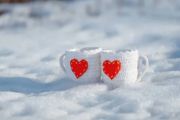 Thermos and cups that stand in the snow. Winter. Knitted covers for mugs. Red hearts on the circles. Thermos with tea.