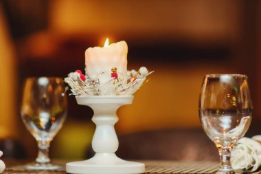 Noel şenlikli tablo ayarı. Kırmızı meyveler ile yanan muma çelenk. Cam şarap gözlük. Bacak beyaz şamdan.