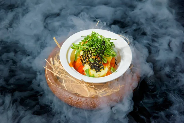 Serving dishes with liquid nitrogen. Salad of vegetables and arugula. Vegetarian dish. Tomatoes and cucumbers. Fresh salad in a white ceramic plate. Dish on a black background