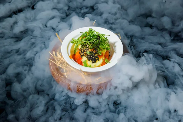Serving dishes with liquid nitrogen. Salad of vegetables and arugula. Vegetarian dish. Tomatoes and cucumbers. Fresh salad in a white ceramic plate. Dish on a black background
