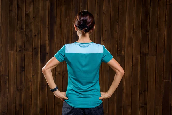 Girl in uniform. The wall is lined with wood. The girl stands against a wooden wall. Blue t-shirt. Fitness watch on the girl's hand.