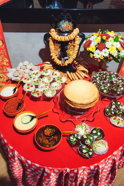 Table setting in Russian traditions. Russian wooden toy in the form of a painted doll. Wooden painted Cutlery. Bouquets of fresh flowers. Drying and gingerbread. Red tablecloth. Khokhloma painting.