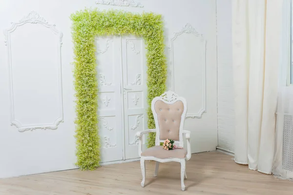 Spring decor. White walls. Beautiful stucco on the walls. Arch of green and white flowers. The decor of the doorway with flowers. White baby\'s breath. The chair has a pale pink color. Wedding bouquet.
