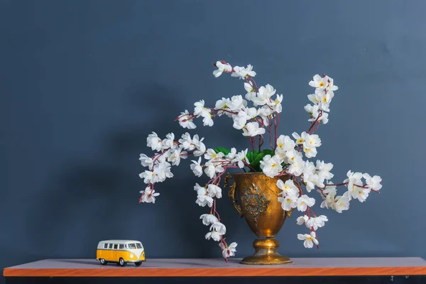 Spring decor. The walls are blue. White flowers in a bronze vase. The interior of the room. Wooden shelf. Decorative element. Toy in the form of a yellow car.