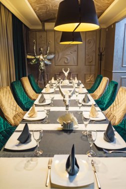 Keçi kafatası heykelcik. Restoranda masa ayarı. Masada gözlük ve plakalar. Çatal bıçak takımı ve Kağıt peçeteler. Siyah avizeler. Sandalyeler altın ve koyu yeşil. Gri masa örtüsü.