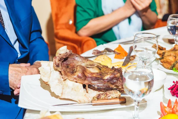 Traditional Kazakh dish of lamb. Roast lamb. Meat dish. Festive table setting. Fruits. Table decor with fresh flowers. Guests at the table. Plates. Tableware.