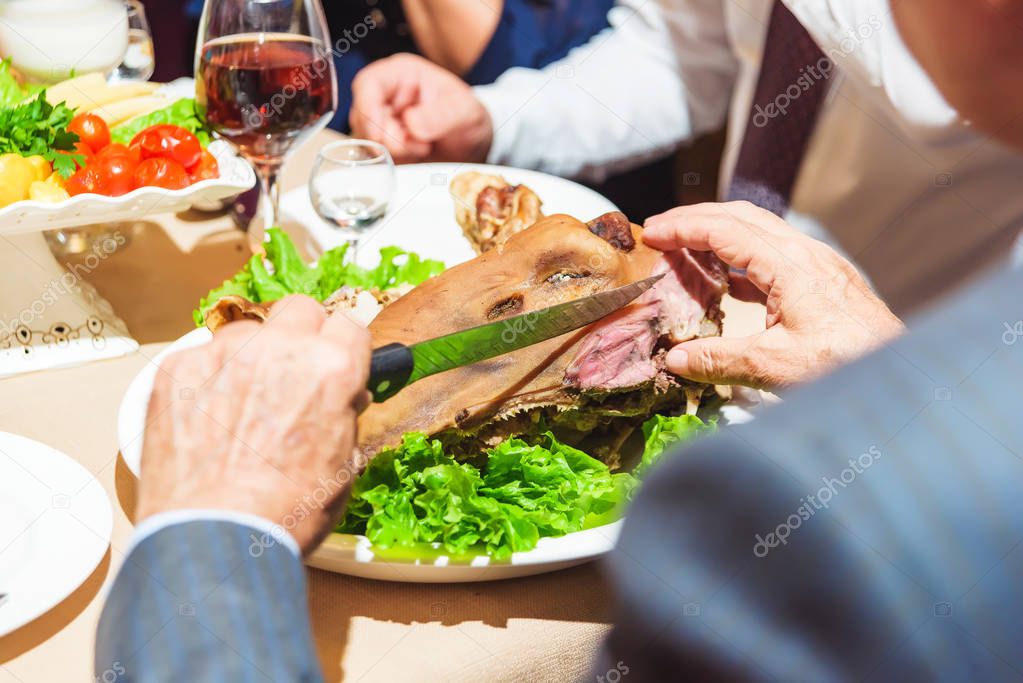 A man cuts a plate of roasted lamb head. Festive table setting. Snacks in plates. Wine glass with a drink on the table.