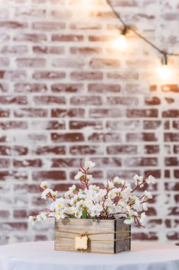 Ahşap kutuda beyaz çiçekler. Tablo beyaz masa örtüsü ile kaplıdır. Tuğla duvar dokusu. Odanın iç mekanı. Duvara asılı ışıklar. Dekoratif tuğla duvar.
