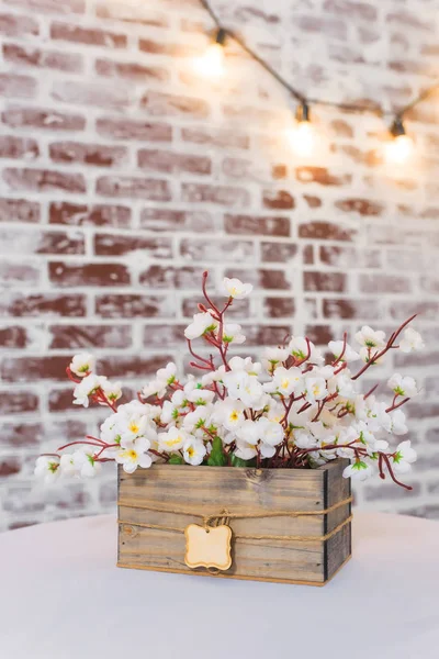 White flowers in a wooden box. Table covered with white tablecloth. Brick wall texture. The interior of the room. Lights on the wire hanging on the wall. Decorative brick wall.