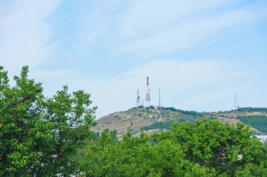 Tepe üzerindeki antenlerle metal yapılar. Tepelik bir arka planda yeşil yaprakları ile ağaçlar. İletişim kulesinde anten.