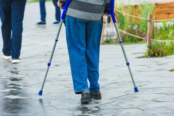 Old grandpa is walking on crutches. Disabled man walking around the city. Metal crutches are blue.