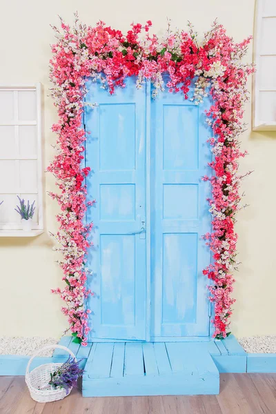 Bahar dekoru. Mavi renkte boyalı ahşap kapılar. Pembe çiçekler ile dekore edilmiş kapılar. Beyaz hasır sepeti. Windows taklit. Tencerelerde lavanta.
