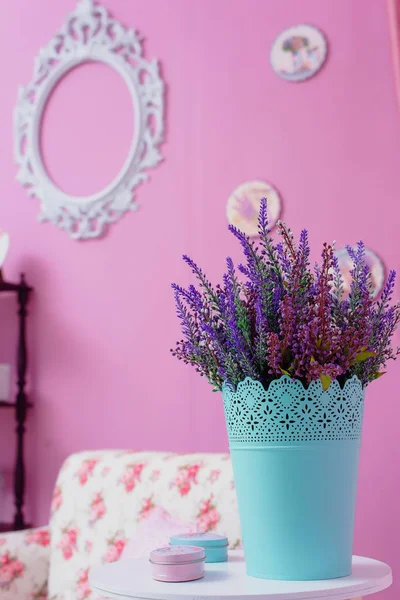 The wall is lilac. Wooden floor shelf.  Decorative element. Lavender in a blue pot. White round table. Pink curtains. Wooden panels on the wall. White chair with floral print.
