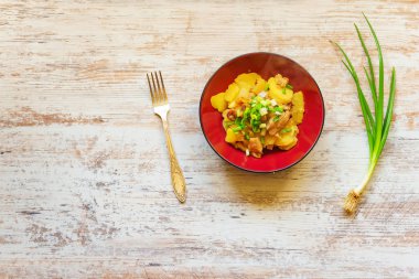 Ahşap masada kırmızı bir tabak kızarmış patates. Bir avuç yeşil soğan ve bir çatal. Lezzetli öğle yemeği.