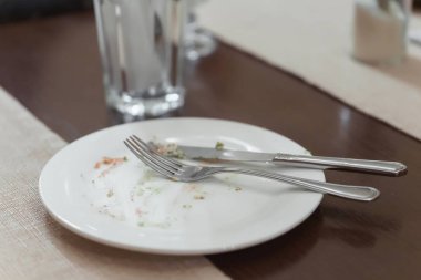 Tahta bir masanın üzerinde kirli bir tatlı tabağı. Çatal bıçak takımı tabakta. Masada dekoratif bej peçeteler.