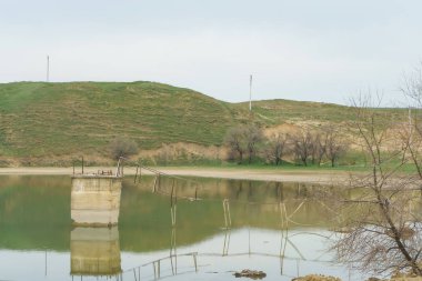 Yeşil tepelerin arka planı üzerinde küçük bir göl. Göldeki eski kırık demir köprü. Su yüzeyinin dokusu.