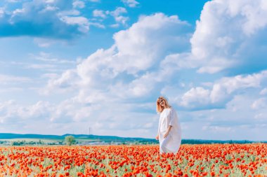 Haşhaş tarlasında beyaz elbiseli bir kız. Kırmızı çiçeklerle tarlada genç bir kadın. Büyük beyaz bulutların arka planında haşhaş tarlasında bir kız.