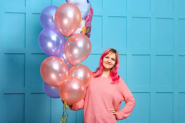 Girl with pink hair with balloons in their hands. Decorating a birthday party in the style of a unicorn. The girl in pink with balloons on a blue background.