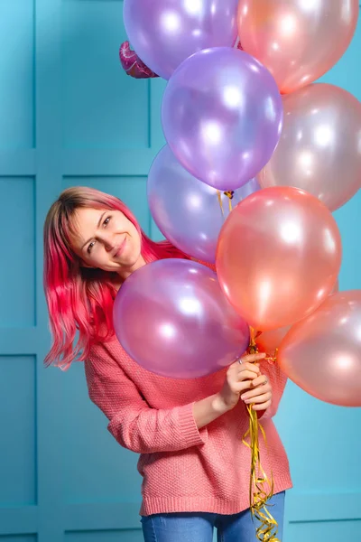 Girl with pink hair with balloons in their hands. Decorating a birthday party in the style of a unicorn. The girl in pink with balloons on a blue background.