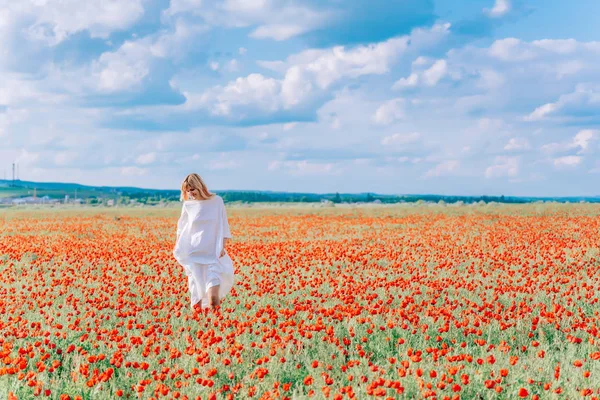 A girl in a white dress in a poppy field. Young woman in the field with red flowers. A girl in a poppy field against the backdrop of large white clouds.