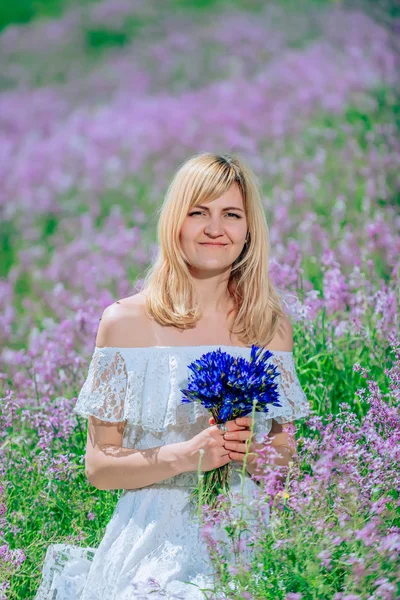 A girl in a white dress on a background of lilac flowers. Girl in a field with a bouquet of cornflowers. Portrait of a Slavic woman with a bouquet of blue flowers in a flowered garden.